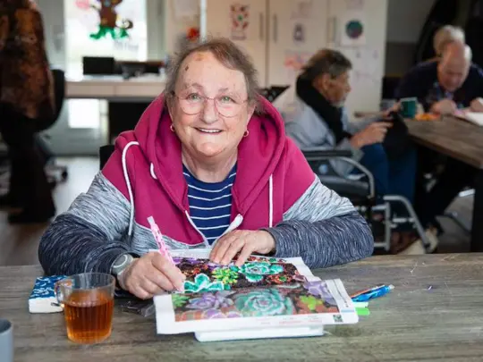 Een oudere vrouw zit te kleuren en lacht in de camera