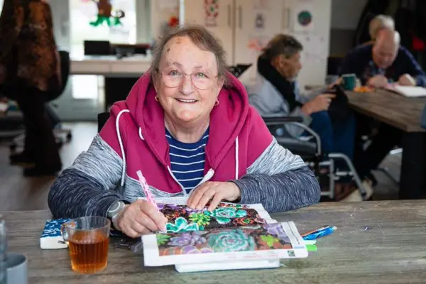 Een oudere vrouw zit te kleuren en lacht in de camera
